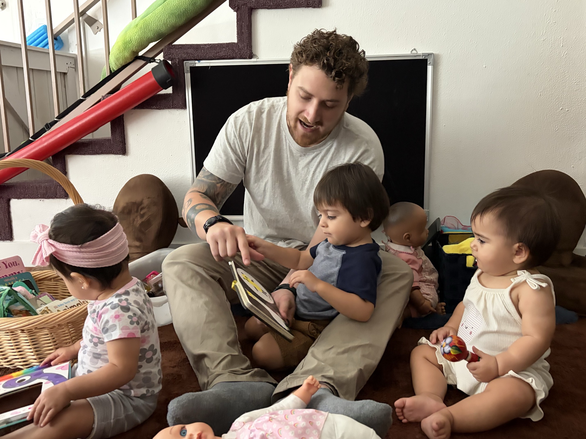 Matthew McArthur sits on a rug reading a board book with three young children; one toddler reaches for the page he is pointing to.